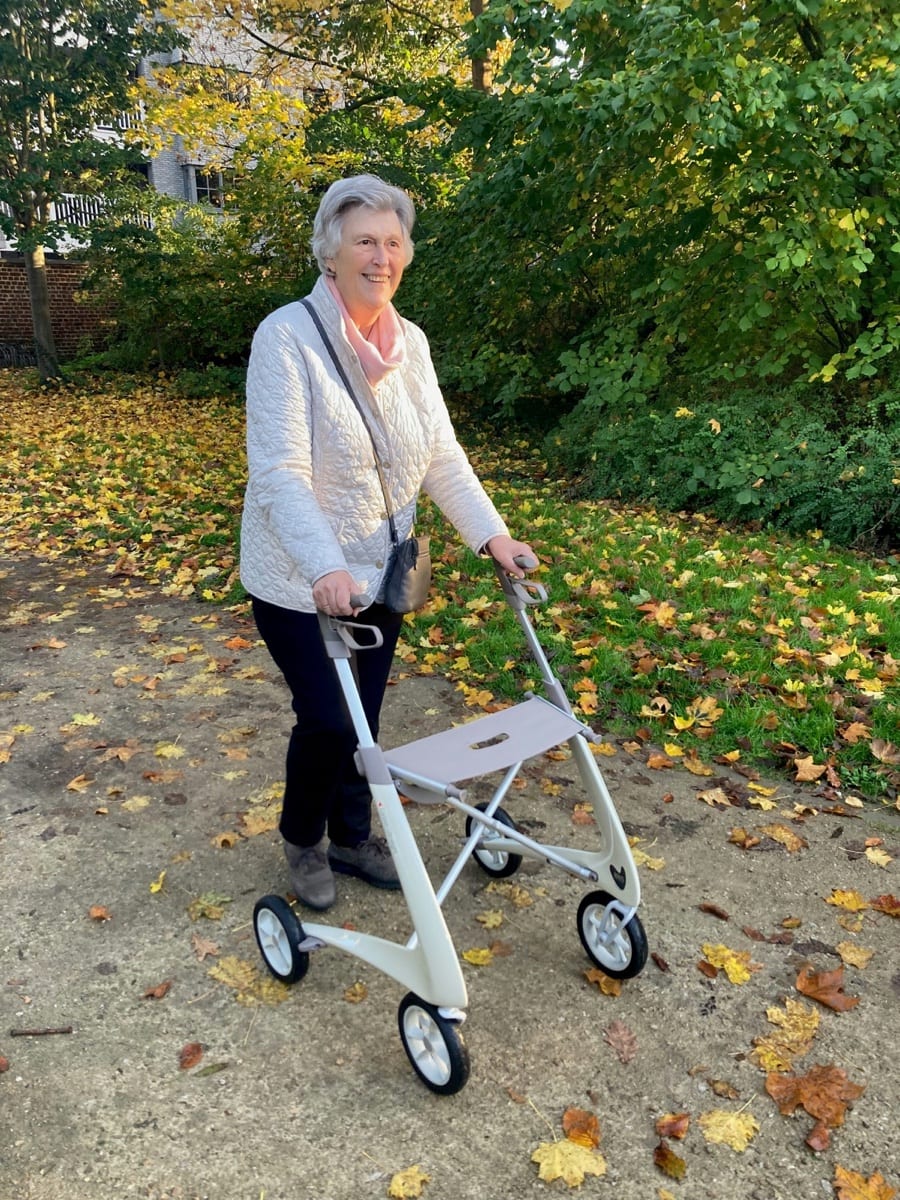 An older adult smiling while using a walker on a leaf-covered outdoor path
